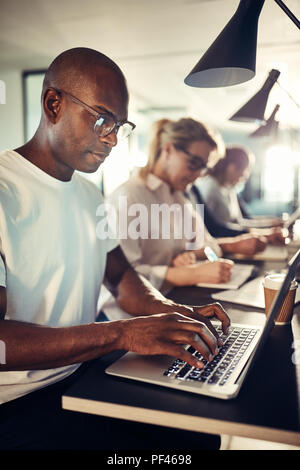 Giovani designer africani incentrata sul suo lavoro mentre è seduto in una riga in una tabella con i colleghi in un ufficio moderno Foto Stock