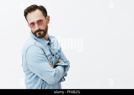 Sei tu che parli con star. Grave-guardando fiduciosi uomo europeo con la barba nel quartiere alla moda di occhiali da sole e giacca denim, tenendo le mani incrociate sul petto mentre in piedi di profilo e fissando sul serio a fotocamera Foto Stock