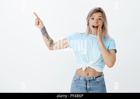 Studio shot di stupito bella donna con tatuaggi e forato ventre, in piedi sconvolto e impressionato, sorridente ampiamente da emozione mentre sta puntando ad angolo superiore sinistro con il dito indice sollevato Foto Stock