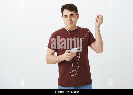Ritratto di orinato scioccato attraente uomo in abito casual, mettendo fuori auricolari con disliking grimace, tenendo lo smartphone, di essere rimasto sorpreso con forte rumore in auricolari oltre il muro grigio Foto Stock