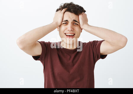 Studio shot di pianto disperato uomo caucasico in t-shirt rossa, toccando i capelli e urlando o sibili, sensazione di miserabili e afflitti, infelice permanente su sfondo grigio Foto Stock