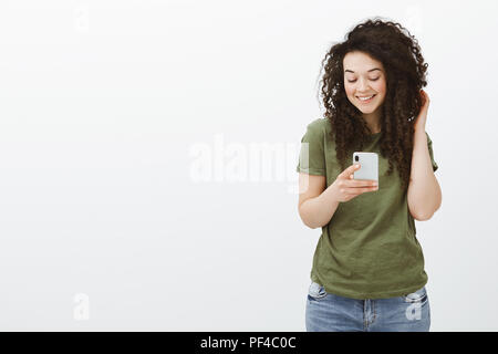 Bella Flirty curly-ragazza dai capelli in verde scuro e t-shirt, toccando i riccioli e sorridente ampiamente con lo schermo dello smartphone durante la visione di un video divertente sul dispositivo o di messaggistica con il mio ragazzo oltre il muro grigio Foto Stock