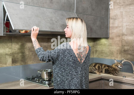 La donna in cucina si apre la casella sospesi fino a quando il gatto in background beve l'acqua dal rubinetto Foto Stock