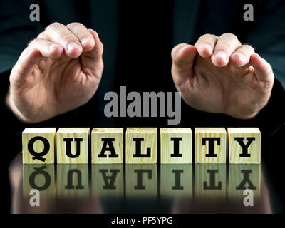 La parola - Qualità - su cubi di legno nero a superficie riflettente con le mani di un imprenditore in un vestito tenuto protettivo sopra i blocchi shelte Foto Stock
