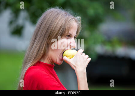 Ritratto di profilo di moody sconvolto arrabbiato attraente bionda con i capelli lunghi giovane donna morde big Apple giallo su sfocata blu verde all'aperto estate backgroun Foto Stock