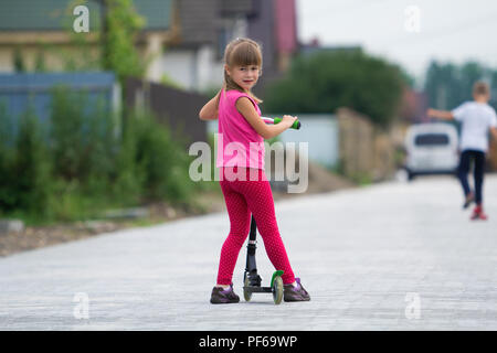 Piuttosto giovane dai lunghi capelli biondi ragazza bambino in rosa vestiti con gli scooter sul sobborgo di sole street e ragazzo su skateboard sfocato estate sfondo luminoso. Foto Stock