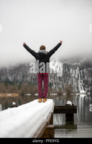 Vista da dietro di un uomo solleva le braccia in trionfo in aria in alto in piedi alla fine di un vecchio molo in legno ricoperta di neve su un tranquillo lago. Foto Stock