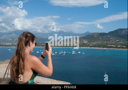 Una giovane donna ventenne prende la foto di una vista sul porto con il suo telefono in Calvi, la Balagne, Corsica, Francia, Europa Foto Stock