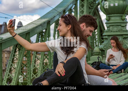 Coppia giovane tenendo selfie sul famoso Ponte della Libertà,noto come libertà,o anche ponte verde di Budapest,lettura giovane ragazza sullo sfondo Foto Stock