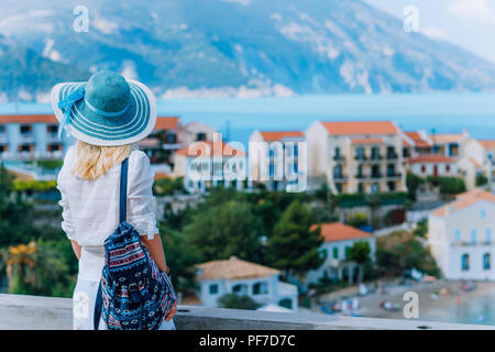 Giovani viaggiatori eleganti donna godendo della vista colorate Assos tranquillo villaggio. Modello femminile che indossa cappello blu, bianco vestiti e zaino di viaggio godendo le vacanze estive a Cefalonia, Grecia Foto Stock