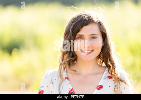 Closeup Ritratto di giovane volto di donna sorridente felice nella soleggiata estate parco verde al tramonto che mostra denti, capelli mossi nel vento, sfondo bokeh di fondo Foto Stock