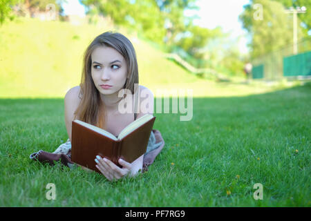 Giovane donna giace su erba verde e legge libro nel parco della città. Foto Stock