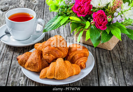 Il francese a forma di mezzaluna rotoli - croissant sul piatto bianco, una tazza di tè e bouquet di fiori freschi e un dono avvolto con della carta su vecchie tavole di legno, Foto Stock