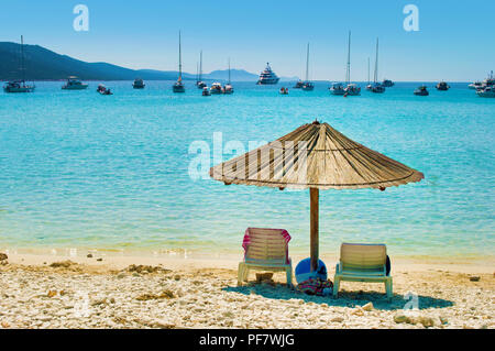 Uno giallo paglierino con ombrellone e due sedie a sdraio sulla sabbia ghiaiosa spiaggia. Molti yacht nel blu mare azzurro laguna in una baia vicino alla costa di Isola. Blu Foto Stock