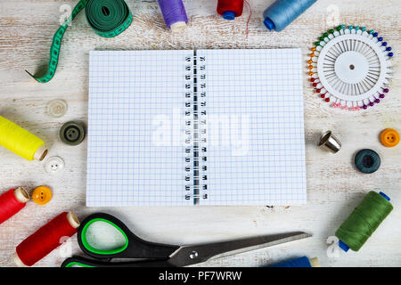 Accessori per il cucito e un notebook aperto su un sfondo di legno. Nastro di misurazione, perni, pulsanti, zipper, forbici e thread. Foto Stock