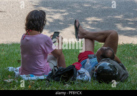 Il marito e la moglie seduta sul prato di un parco godendo il sole e utilizzando un telefono cellulare o un dispositivo portatile. Argento surfisti utilizzando la tecnologia. Foto Stock