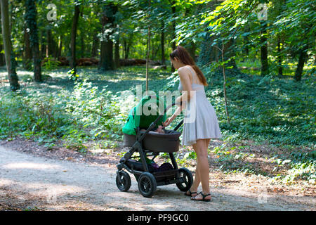 Passeggiata nella foresta con passeggino, vista laterale del giovane mamma nel bel vestito camminando sul percorso con il suo bambino nel passeggino, godendo di aria fresca Foto Stock