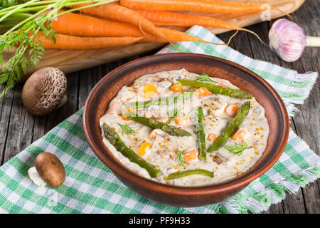 Delizioso formaggio cremosa zuppa di funghi con molla carote, gli asparagi e le spezie, decorata con aneto, in un vaso di argilla su un vecchio tavolo rustico, horizonta Foto Stock