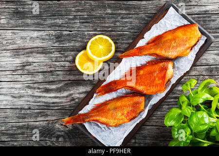 Red Sea Bass su una carta pergamena su un vecchio tavolo di legno con foglie di basilico e fettine di limone sullo sfondo, vista dall'alto Foto Stock