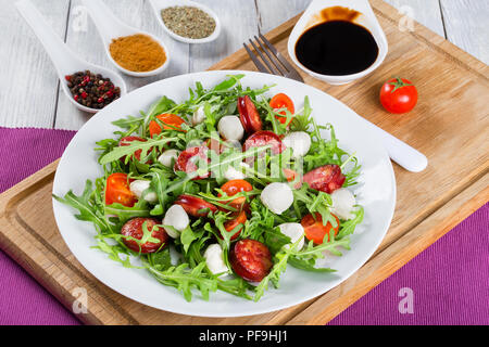 Delizioso con insalata di pomodori, mini mozzarella, rucola, salsicce fritte su un piatto bianco su bianco tavolo rustico, caramellato aceto balsamico in un gra Foto Stock