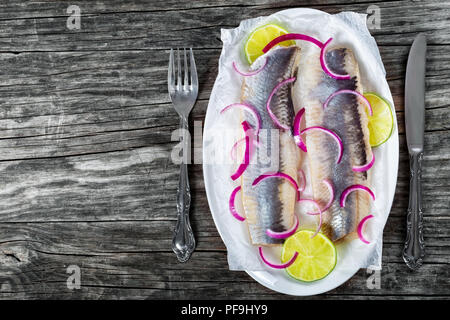 Filetti di aringhe con cipolla e fettine di lime su carta pergamena su un ovale piatto su un vecchio tavolo rustico con la lama di un coltello e forchetta, vista dall'alto Foto Stock