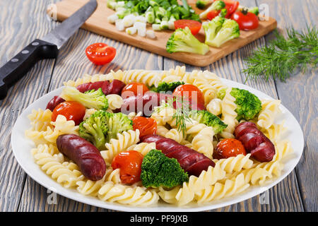 Spirale deliziosa insalata di pasta con broccoli e salsicce alla griglia su un piatto bianco con pomodoro fresco e sorrel insalata verde, cipolline su un taglio Foto Stock