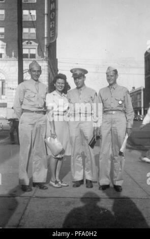 Fotografia in bianco e nero che mostra un sorridente giovane donna, che indossa un mantello e tenendo un grande frizione, in piedi tra tre sorridente uomini vestiti in uniformi militari, tutti in piedi su una strada di città angolo, in piena vista di lunghezza, fotografato in Ohio, 1945. () Foto Stock