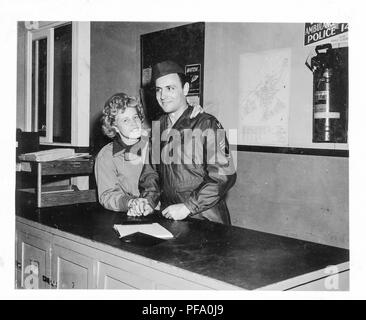 Fotografia in bianco e nero, che mostra un uomo in piedi accanto a una bionda donna, entrambi mostrati dalla vita in su, indossano uniformi militari e di stare al chiuso, dietro un lungo bancone, con un estintore in background, l'uomo sorrisi a qualcosa al di fuori della telecamera mentre la donna si affaccia la telecamera, loro legato le mani a destra il resto sul bancone mentre la donna di braccio sinistro avvolge l'uomo della spalla, probabile fotografato durante la II Guerra Mondiale, 1945. () Foto Stock