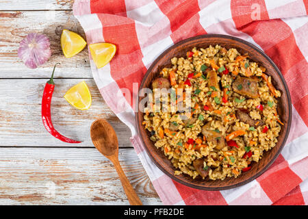 Riso con carne, pepe, spezie e verdure sul piatto, fetta di limone, aglio, peperoncino e cucchiaio di legno sulle bianche pareti scrostate plance, vista da abo Foto Stock