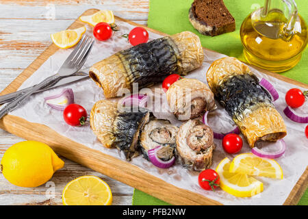 Un sano cotto i filetti di sgombro in rotoli, i pomodori e le fette di limone su carta pergamena bianca sul tagliere con coltello e forchetta,cipolla e bottiglia di Foto Stock