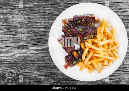 Fritto delizioso nervature appiccicosa cosparsi di cipolla verde con patatine fritte sul piatto bianco sul vecchio di legno scuro dello sfondo, vista da sopra Foto Stock