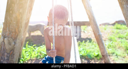 Banner sfocatura dello European ragazzo seduto su una tavola di legno vecchio oscillare in un parco dal mare. Vacanze in villaggio giochi nella natura Foto Stock