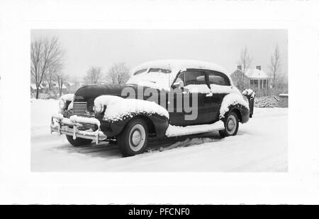 Fotografia in bianco e nero, mostrando una tre-quarto vista di profilo di un oscuro, vintage, Chevrolet sedan auto parcheggiate fuori in un paesaggio wintery, di fresco con neve caduta sul tetto, modanature, parabrezza e l'esecuzione di bordo, con alberi sfrondato e case in background, probabilmente fotografato in Ohio nel decennio dopo la II Guerra Mondiale, 1945. () Foto Stock