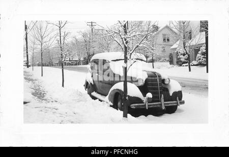 Fotografia in bianco e nero, mostrando una leggermente angolata vista frontale di un oscuro, vintage, Chevrolet sedan auto parcheggiate fuori in un paesaggio wintery, di fresco con neve caduta sul tetto, modanature, parabrezza e l'esecuzione di bordo, con una struttura ad albero sfrondato in primo piano e altri alberi e case in background, probabilmente fotografato in Ohio nel decennio dopo la II Guerra Mondiale, 1945. () Foto Stock