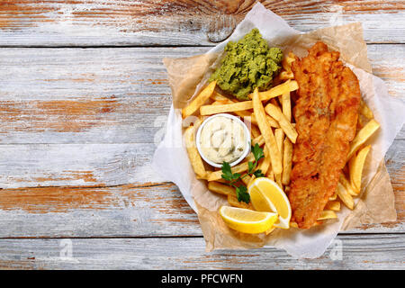 Delizioso croccante di pesce e patatine fritte - cod, patatine fritte, le fette di limone, salsa tartara e purea di piselli su piastra su carta sul vecchio tavolo in legno, authen Foto Stock