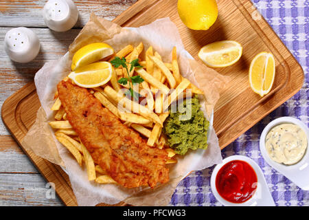 Croccante di pesce e patatine fritte - cod, patatine fritte, le fette di limone, salsa tartara e purea di piselli sulla piastra su carta sul vecchio tavolo in legno con rosmarino in mor Foto Stock