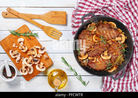 Delizioso filetto di maiale in umido con cipolla. funghi, rosmarino, semi di vino bianco secco e spezie in stufato di ferro coppa sul tavolo bianco con spatole in legno, facile Foto Stock