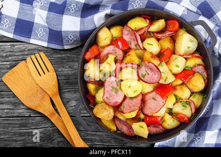 Gustoso arrosto Spicchi di patate con prosciutto e peperone rosso, spruzzata con fresco verde primavera la cipolla nella padella di ferro sul buio tavolo rustico con carta asciugatutto, Foto Stock