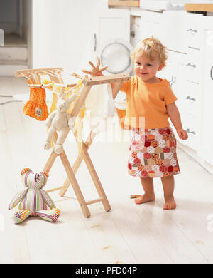 Bionda sorridente toddler in giallo T-shirt e grembiule in piedi in cucina appendere i vestiti e giocattoli morbidi sulla linea di lavaggio, vista frontale. Foto Stock