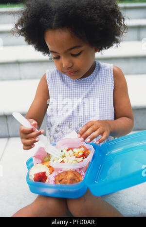 La ragazza con i capelli ricci seduti sui passi all'aperto e mangiare da una scatola di pranzo sul suo grembo, vista frontale. Foto Stock