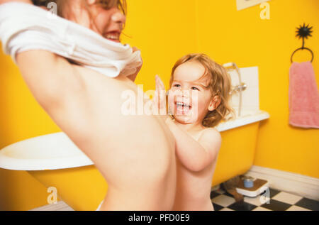 Due ragazze in piedi nel bagno di colore giallo con pavimento a scacchi, uno stiro indietro mettendo sulla sua sommità, l'altra giovane ragazza ridere per lei. Foto Stock