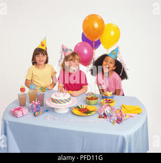 Tre ragazze in parte cappelli a sorridere e ridere mentre è seduto a tavola decorata per la festa di compleanno, con cibo e bevande e palloncini, due ragazze guardando la fotocamera. Foto Stock
