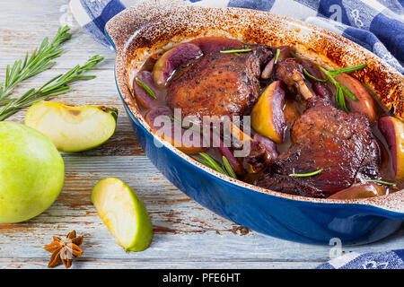 Grigliata di gambe di anatra cotto al forno con vino rosso e rosmarino,la scorza di arancia e fette di mela in posizione gratin del piatto sulla tovaglia, mela verde su bianco peeling pa Foto Stock