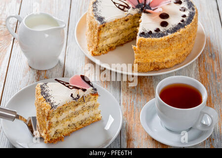 Pan di Spagna con semi di papavero a strati con la crema con un taglio fuori pezzo sulla torta la pala sulla piastra bianca di close-up con brocca del latte, la tazza di tè nero su sa Foto Stock