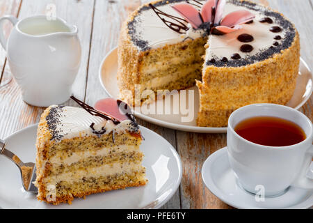 Pan di Spagna con semi di papavero a strati con la crema con un taglio fuori pezzo sulla torta la pala sulla piastra bianca di close-up con brocca del latte, la tazza di tè nero su sa Foto Stock