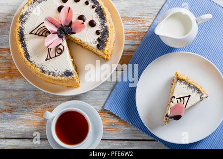 Pan di Spagna con semi di papavero a strati con la crema con un taglio fuori pezzo sulla piastra bianca close-up sulla tabella MAT con la brocca del latte, la tazza di tè nero su sauc Foto Stock