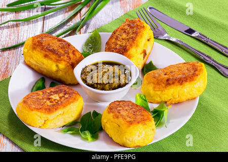 Deliziose frittelle di patate da purea di patate con salsa di pesto e foglie di basilico bianco sul piatto sul tavolo verde mat, verde cipollina sul retro Foto Stock