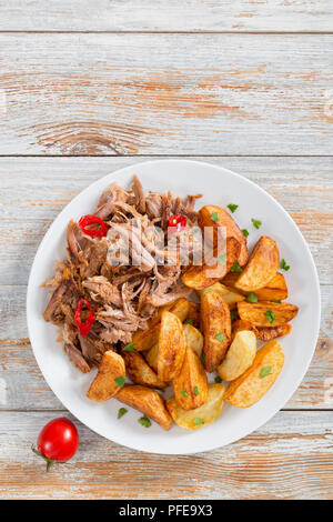 Porzione di tirata slow-cotta deliziosa carne arrostita al forno con patate fritte cunei cosparsa di prezzemolo sulla piastra bianca con spezie e peperoncino su Foto Stock