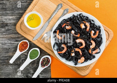 Deliziosa seppia tagliatelle nere con gamberoni sul piatto bianco sul tagliere con olio d'oliva e spezie in cucchiai sullo sfondo, vista da sopra