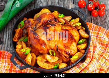 Dorate e croccanti della pelle di pollo grigliato in forno con spicchi di patate e porri in ghisa pan su vecchie tavole di legno, tagliere con fette di cipolla e Foto Stock